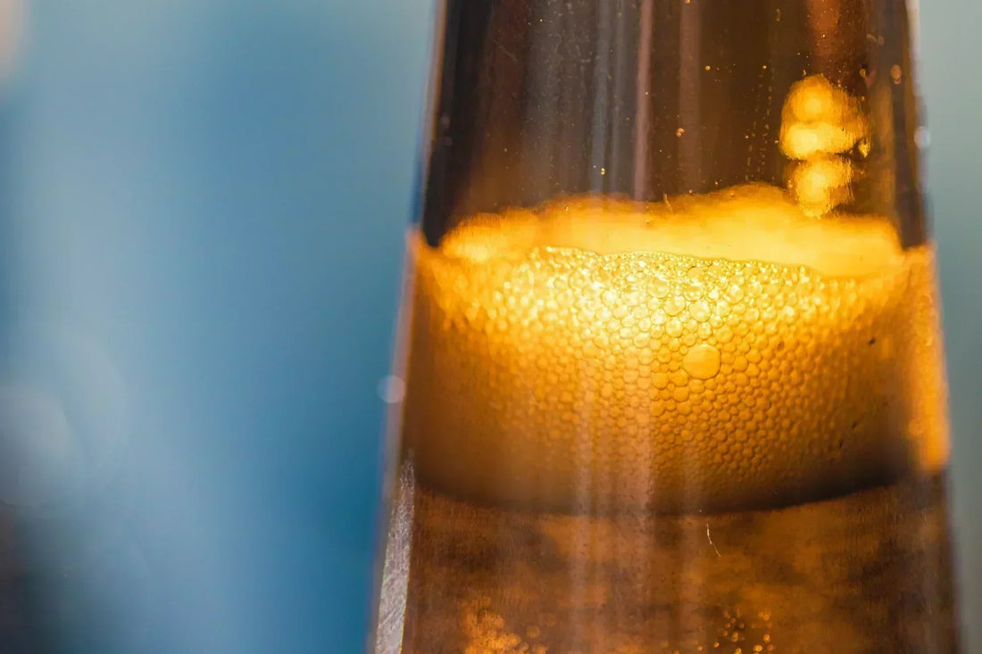 Bouteille de kombucha Galatea versée dans un verre, lumière chaude et dorée, bulles en suspension, évoquant une transformation alchimique de la matière en vie