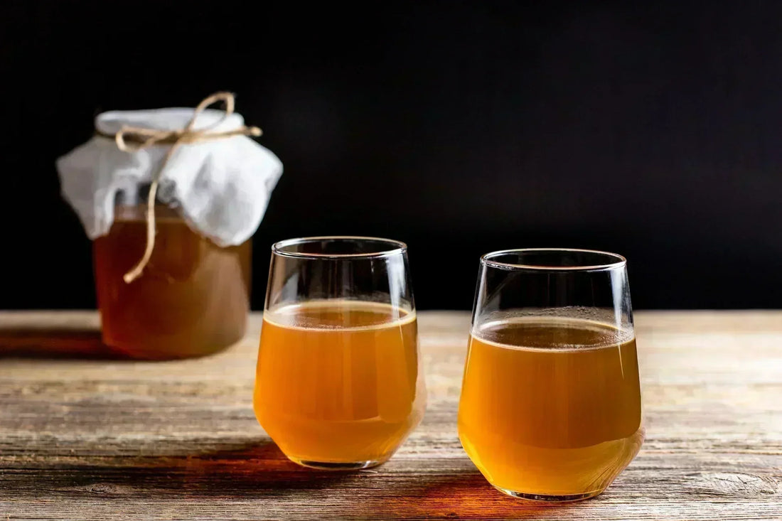 Verre de kombucha ambré aux bulles fines, posé près d'une jarre sur une table en bois
